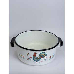 May include: White enamelware pot with a black rim and handles. The pot features a colorful rooster design with floral and house motifs. The pot is a classic kitchenware item.