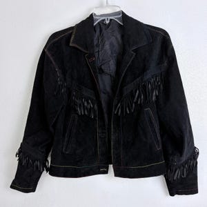 May include: A black suede jacket featuring long fringe detailing across the chest and sleeves. The jacket includes a classic collar, a button-front closure, two vertical front pockets, and visible contrast stitching along the seams.