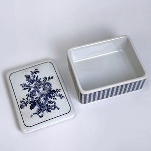 May include: A rectangular ceramic box with a removable lid. The lid features a blue floral bouquet illustration on a white background. The exterior sides of the box are decorated with a blue and white plaid pattern, while the interior is plain white.
