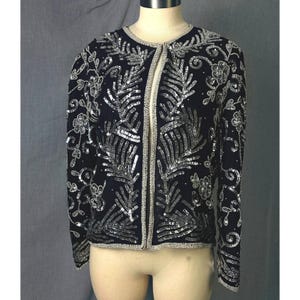 May include: A black jacket with silver sequin and bead embellishments. The jacket features a round neckline and a straight, open front. The design includes floral and leaf patterns, with silver trim outlining the edges. The jacket is displayed on a mannequin.