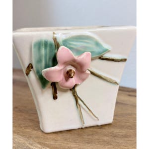 May include: A square-shaped ceramic planter with a glossy off-white finish. The front features a raised, three-dimensional pink flower with a brown center, accompanied by painted green leaves and brown stems extending across the surface.