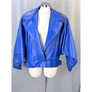 May include: A vibrant blue leather jacket with a wide collar and textured shoulder and lapel accents. The jacket features a button closure and a belted waist, displayed on a mannequin. The sleeves are wide and the overall style is vintage.