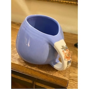May include: A pale blue ceramic mug with a distinctive, rounded form. The handle is white with a floral pattern in orange, pink, and green. The mug is on a wooden surface.
