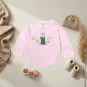 K&ouml;nnte beinhalten: Ein hellrosa Baby-Body mit langen &Auml;rmeln. Auf der Vorderseite befindet sich eine gr&uuml;n-wei&szlig;e Grafik eines Guinness-Pints, das von zwei H&auml;nden gehalten wird, mit einer Krone dar&uuml;ber. Eine beige M&uuml;tze, gestrickte Babyschuhe und Holzspielzeug sind ebenfalls zu sehen.