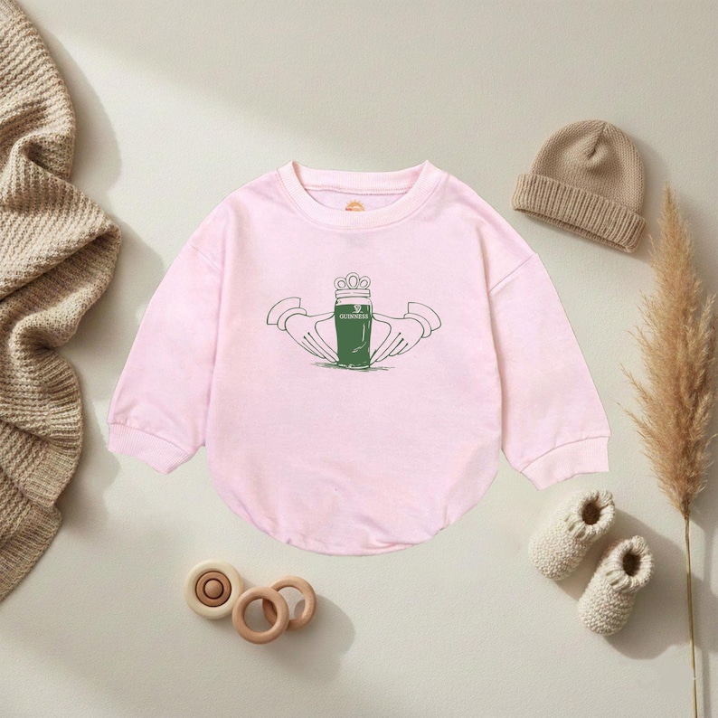 K&ouml;nnte beinhalten: Ein hellrosa Baby-Body mit langen &Auml;rmeln. Auf der Vorderseite befindet sich eine gr&uuml;n-wei&szlig;e Grafik eines Guinness-Pints, das von zwei H&auml;nden gehalten wird, mit einer Krone dar&uuml;ber. Eine beige M&uuml;tze, gestrickte Babyschuhe und Holzspielzeug sind ebenfalls zu sehen.