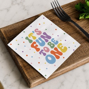 May include: White cocktail napkin with the text "IT'S FUN TO BE ONE" in colorful, retro-style lettering. The napkin is decorated with small, colorful polka dots and rests on a wooden cutting board with a black fork.