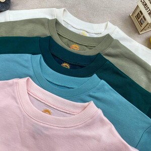 K&ouml;nnte beinhalten: Ein Stapel Rundhals-Sweatshirts in verschiedenen Farben: Wei&szlig;, Olivgr&uuml;n, T&uuml;rkis, Hellblau und Rosa. Jedes Sweatshirt hat einen gerippten Ausschnitt und ein kleines gesticktes Sonnenlogo. Die Sweatshirts sind ordentlich angeordnet und zeigen ihre verschiedenen Farbt&ouml;ne.