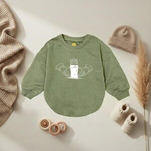 K&ouml;nnte beinhalten: Salbeigr&uuml;nes Baby-Sweatshirt mit wei&szlig;er Grafik eines Guinness-Pints mit Krone und H&auml;nden. Eine beige Strickm&uuml;tze, beige Babyschuhe und Holzspielzeug sind um das Sweatshirt angeordnet.