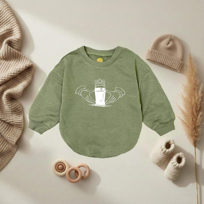K&ouml;nnte beinhalten: Salbeigr&uuml;nes Baby-Sweatshirt mit wei&szlig;er Grafik eines Guinness-Pints mit Krone und H&auml;nden. Eine beige Strickm&uuml;tze, beige Babyschuhe und Holzspielzeug sind um das Sweatshirt angeordnet.
