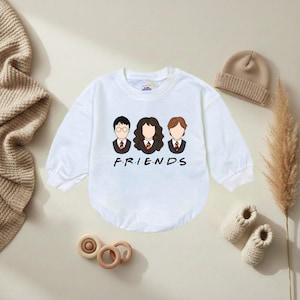 May include: White long-sleeved baby bodysuit with a cartoon graphic of three characters and the word "FRIENDS" in black. The characters are wearing ties and blazers. A beige beanie, knitted booties, and wooden toys are also in the image.