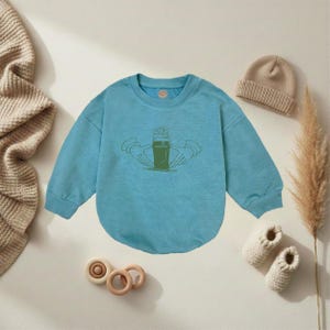 K&ouml;nnte beinhalten: Hellblaues Baby-Sweatshirt mit gr&uuml;ner Grafik eines Bierglases, das von H&auml;nden gehalten wird. Ein beigefarbener Strickhut, ein Paar wei&szlig;e Strick-Babyschuhe und Holzspielzeug sind ebenfalls im Bild.
