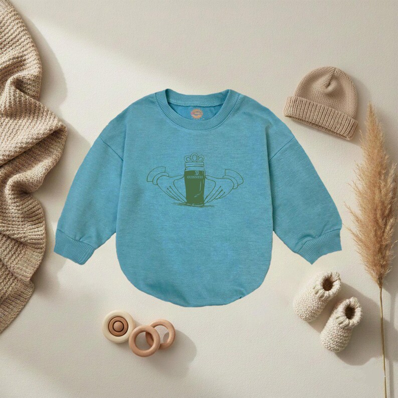 K&ouml;nnte beinhalten: Hellblaues Baby-Sweatshirt mit gr&uuml;ner Grafik eines Bierglases, das von H&auml;nden gehalten wird. Ein beigefarbener Strickhut, ein Paar wei&szlig;e Strick-Babyschuhe und Holzspielzeug sind ebenfalls im Bild.