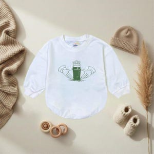 K&ouml;nnte beinhalten: Wei&szlig;er Baby-Body mit langen &Auml;rmeln und gr&uuml;nem Claddagh-Design. Der Claddagh zeigt eine Krone, H&auml;nde und ein herzf&ouml;rmiges Objekt. Ein beigefarbener Strickhut, ein Paar gestrickte Babyschuhe und Holzspielzeug sind ebenfalls im Bild.