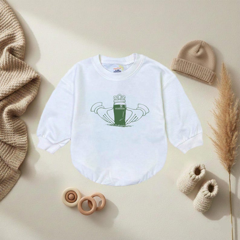 K&ouml;nnte beinhalten: Wei&szlig;er Baby-Body mit langen &Auml;rmeln und gr&uuml;nem Claddagh-Design. Der Claddagh zeigt eine Krone, H&auml;nde und ein herzf&ouml;rmiges Objekt. Ein beigefarbener Strickhut, ein Paar gestrickte Babyschuhe und Holzspielzeug sind ebenfalls im Bild.