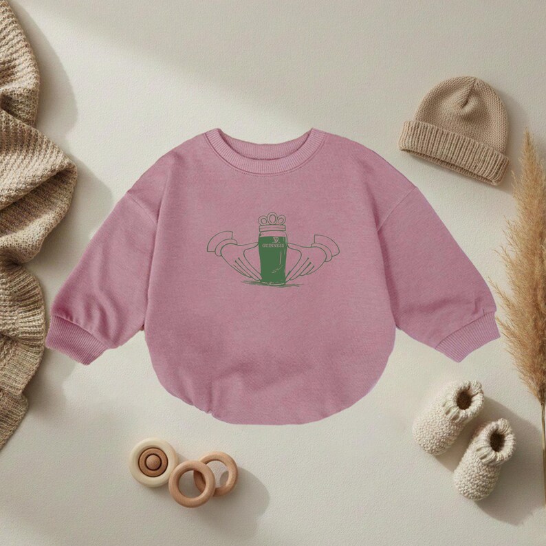 K&ouml;nnte beinhalten: Ein hellrosa Baby-Sweatshirt mit einem gr&uuml;nen Claddagh-Design. Das Claddagh-Symbol mit Krone, H&auml;nden und Herz steht f&uuml;r Liebe, Loyalit&auml;t und Freundschaft. Das Sweatshirt hat lange &Auml;rmel und einen runden Halsausschnitt. Weitere Artikel: beigefarbene M&uuml;tze, Babyschuhe und Holzspielzeug.