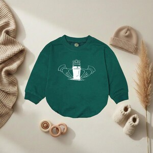 K&ouml;nnte beinhalten: Dunkelgr&uuml;nes Baby-Sweatshirt mit wei&szlig;er Grafik eines Guinness-Pints in einem Claddagh-Ring. Das Sweatshirt hat lange &Auml;rmel und einen Rundhalsausschnitt. Ein beigefarbener Beanie und gestrickte Babyschuhe sind ebenfalls im Bild.