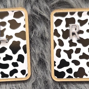 May include: Two rectangular cow print coasters with gold trim. One coaster has a silver "L" and the other has a silver "R". The coasters are set against a gray, fuzzy background.