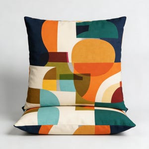 Pode incluir: Duas almofadas quadradas com uma borda azul marinho e um padrão geométrico colorido. O design apresenta formas laranja, verde-azulado, amarelas e verdes sobre um fundo branco sujo. As almofadas estão empilhadas uma sobre a outra.