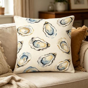 May include: Square cushion featuring a cream background with a repeating watercolour oyster design in blue, grey, and yellow. The cushion is on a beige sofa with other cushions.