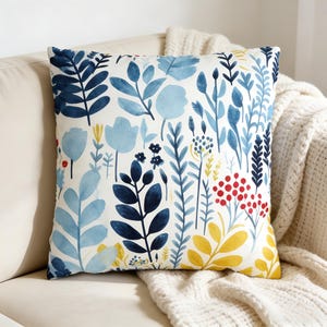 Könnte beinhalten: Quadratisches Zierkissen mit botanischem Design. Das Kissen hat einen weißen Hintergrund mit Aquarell-Illustrationen von blauen, marineblauen, gelben und roten Blumen und Blättern. Das Kissen liegt auf einem beigen Sofa mit einer weißen Decke.