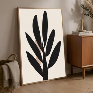 May include: A framed art print featuring a minimalist design. The artwork showcases a black, stylized plant against a cream-colored background. The plant's leaves are elongated and abstract. The frame is a light brown color.