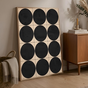 May include: A framed artwork featuring a grid of twelve black circles on a light beige background. The artwork is displayed against a neutral wall, with a wooden cabinet and a woven basket in the background. The frame is a light wood color.