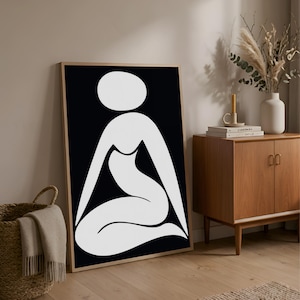 May include: A framed art print featuring a minimalist design of a white figure on a black background. The artwork is displayed in a room with a wooden cabinet, a woven basket, and a beige rug, creating a modern aesthetic.