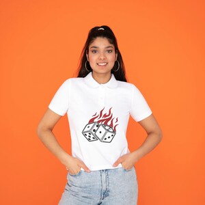 May include: White polo shirt featuring a graphic of two dice engulfed in flames. The shirt has a classic collar and short sleeves. The background is a solid orange color.