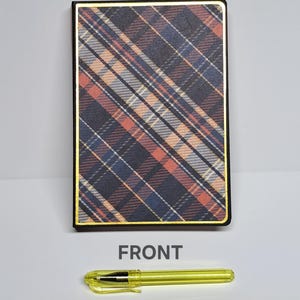 May include: A rectangular notebook with a plaid pattern in shades of brown, blue, and orange, framed with a gold border. A yellow pen is at the bottom, and the word "FRONT" is printed below the notebook.
