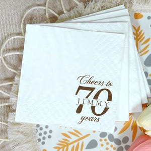 May include: A stack of white paper napkins with a textured border. The top napkin features dark brown printed text that reads Cheers to 70 JIMMY years, with the number 70 prominently displayed in a large, bold, dark brown sans-serif font.