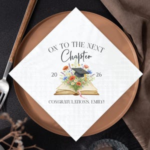 May include: A white square napkin with the text "ON TO THE NEXT Chapter" and "20 26". The napkin features a watercolor illustration of an open book, flowers, and a graduation cap. The text "CONGRATULATIONS, EMILY!" is also present.