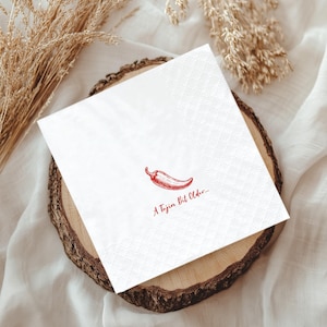 May include: White square paper napkin with a red chili pepper illustration and the text "A Tajin Bit Older...". The napkin is placed on a wooden slice, with dried plants in the background.