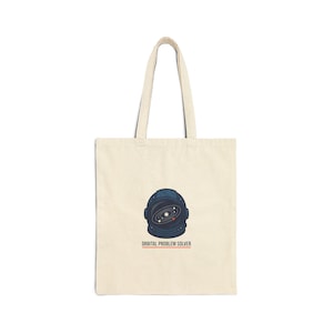 Op de afbeelding: Een crèmekleurige draagtas met een marineblauw astronautenhelm-ontwerp en een galaxy-illustratie. De tekst "ORBITAL PROBLEM SOLVER" is onder de helm in oranje en wit gedrukt. De tas heeft twee lange handvatten.