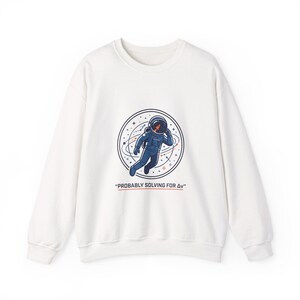 Puede incluir: Sudadera blanca de cuello redondo con un gráfico de un astronauta con un traje espacial azul dentro de un diseño circular. El texto "PROBABLY SOLVING FOR Δv" está debajo. Sudadera de estilo casual.