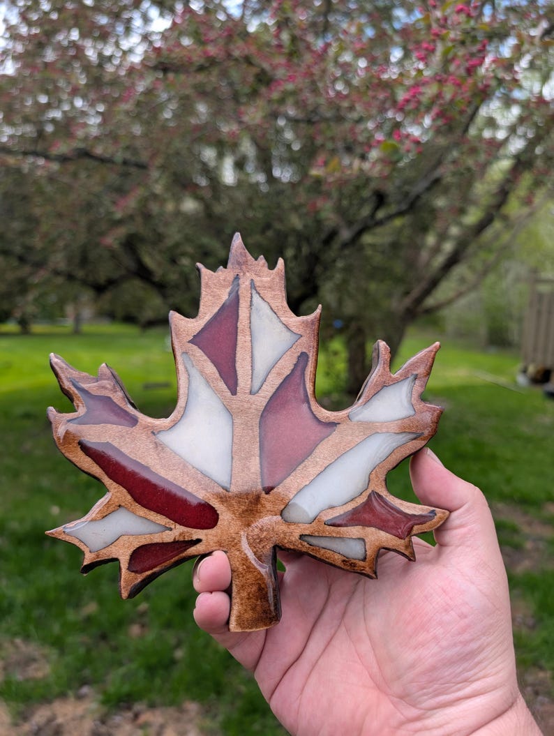 May include: A handcrafted maple leaf ornament with a stained wood border and inlaid sections of red and white. The leaf is held in a hand against a blurred outdoor background.
