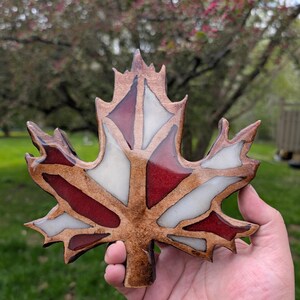 May include: A wooden maple leaf with red, white, and brown sections. The leaf is held in a hand against a blurred background of green grass and a tree with pink blossoms. The leaf is a decorative item.