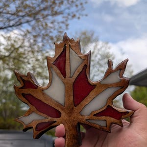 May include: A wooden maple leaf with red and white accents. The leaf is held up against a cloudy sky. The leaf's edges are a dark brown, and the red and white sections are smooth and glossy.