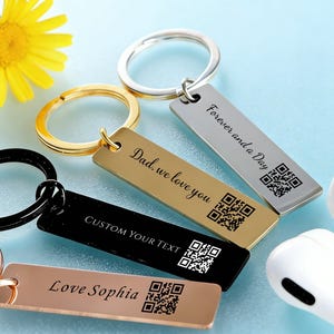 May include: A collection of rectangular keychains in gold, silver, black, and rose gold. Each keychain has a ring and engraved text, including "Forever and a Day," "Dad, we love you," "Custom Your Text," and "Love Sophia." A yellow flower and white earbuds are also in the image.