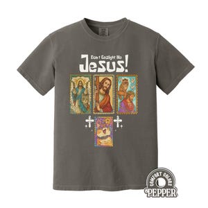 May include: Dark gray t-shirt with the text "Don't Gaslight Me Jesus!" above four postage stamp-style images of Jesus and a cat. The shirt also has the Comfort Colors Pepper logo.