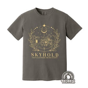 Puede incluir: Una camiseta gris con un diseño dorado de un castillo, montañas y elementos celestiales. El texto "SKYHOLD" y "THE PLACE WHERE THE SKY IS KEPT" están debajo. La camiseta es de la marca Comfort Colors.