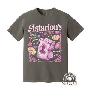 May include: Dark grey t-shirt featuring a pink graphic of a juice box with the text "Astarion's Juice Box". The design includes illustrations of fruit and the words "100% Organic" and "His favorite".