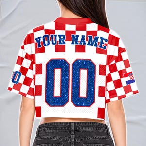 May include: Cropped white and red checkered jersey with the text "YOUR NAME" in blue. The jersey has the number "00" in blue glitter and a Croatian flag on the sleeve. The jersey has a red collar and sleeve trim.