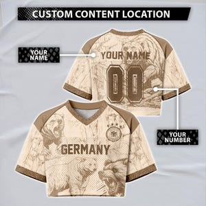 Könnte beinhalten: Ein beiges und braunes bauchfreies Trikot mit V-Ausschnitt, Bärenillustrationen, dem Schriftzug GERMANY und einem runden Emblem. Das Design enthält Platzhalter für YOUR NAME und YOUR NUMBER sowie ein Banner CUSTOM CONTENT LOCATION.