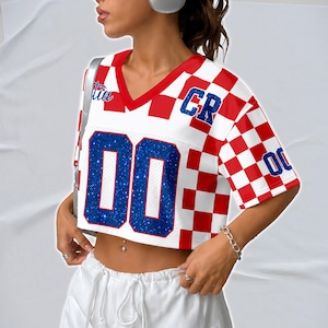 May include: A cropped white and red checkered jersey-style top with blue glitter numbers and lettering. The top has a V-neck and short sleeves. The number "00" is prominently displayed on the front.