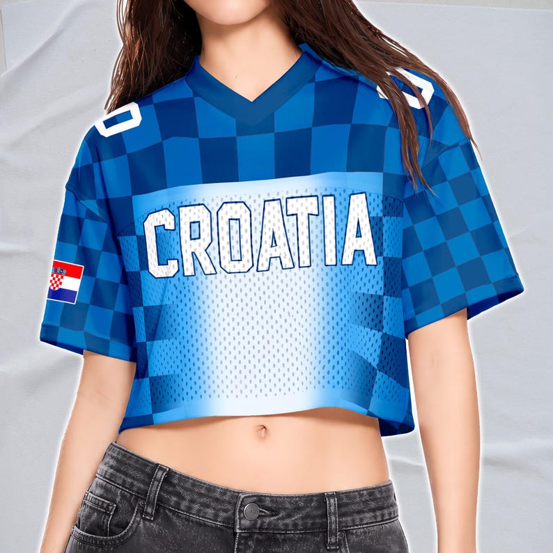 May include: Cropped blue jersey with the word "CROATIA" in white block letters. The jersey has a checkered pattern on the sleeves and shoulders, with the Croatian flag on the left sleeve. The jersey has a V-neck.