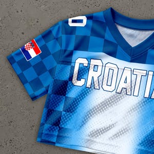 May include: Blue and white Croatia jersey with the word "CROATIA" in large white letters. The jersey features a checkered pattern on the sleeves and a Croatian flag patch. The jersey has a V-neck design.