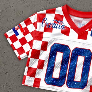 May include: A white and red checkered jersey with the word "Croatia" in blue script. The jersey has blue glitter numbers "00" on the front and a Croatian flag on the sleeve. The jersey has a red V-neck collar.