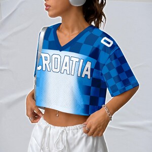 May include: A blue and white crop top with the word "CROATIA" in white block letters. The top has a checkered pattern on the sleeves and shoulders. The shirt has a V-neck and short sleeves.