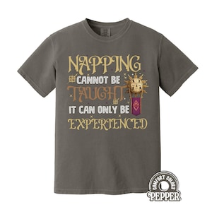 Puede incluir: Camiseta gris con el texto dorado "Napping Cannot Be Taught It Can Only Be Experienced". El diseño incluye un gráfico similar al sol y una pancarta morada. La camiseta tiene mangas cortas y cuello redondo.