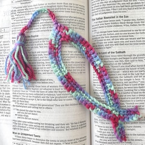 Pode incluir: Um marcador de livros de crochê feito à mão em tons de rosa, roxo, azul e verde. O marcador de livros tem uma borla no topo e está sobre um livro aberto. O marcador de livros tem cerca de 25 cm de comprimento.